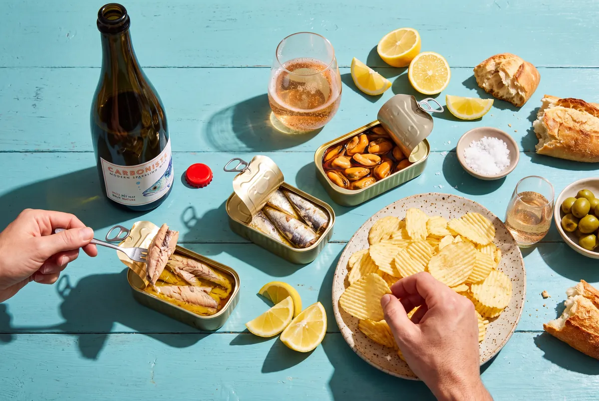 A bottle of 2023 Carboniste Mackerel pét-nat on a sunny blue table with open tins of mackerel, sardines and mussels, a plate of ridged potato chips, lemons and two glasses of copper sparkling wine