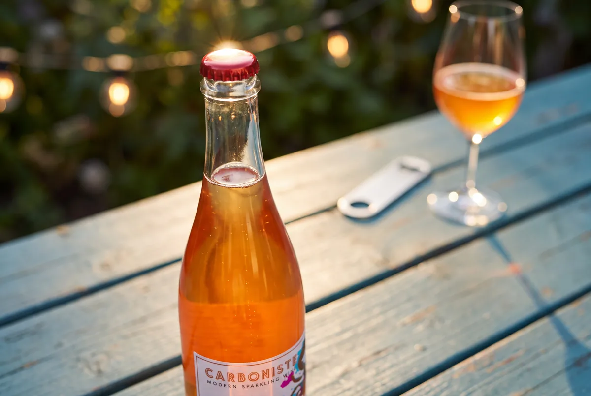 Close-up of the red crown cap on a bottle of 2024 Carboniste Crab sparkling orange wine on a blue picnic table at golden hour, with a bottle opener and a glass of orange sparkling wine behind it