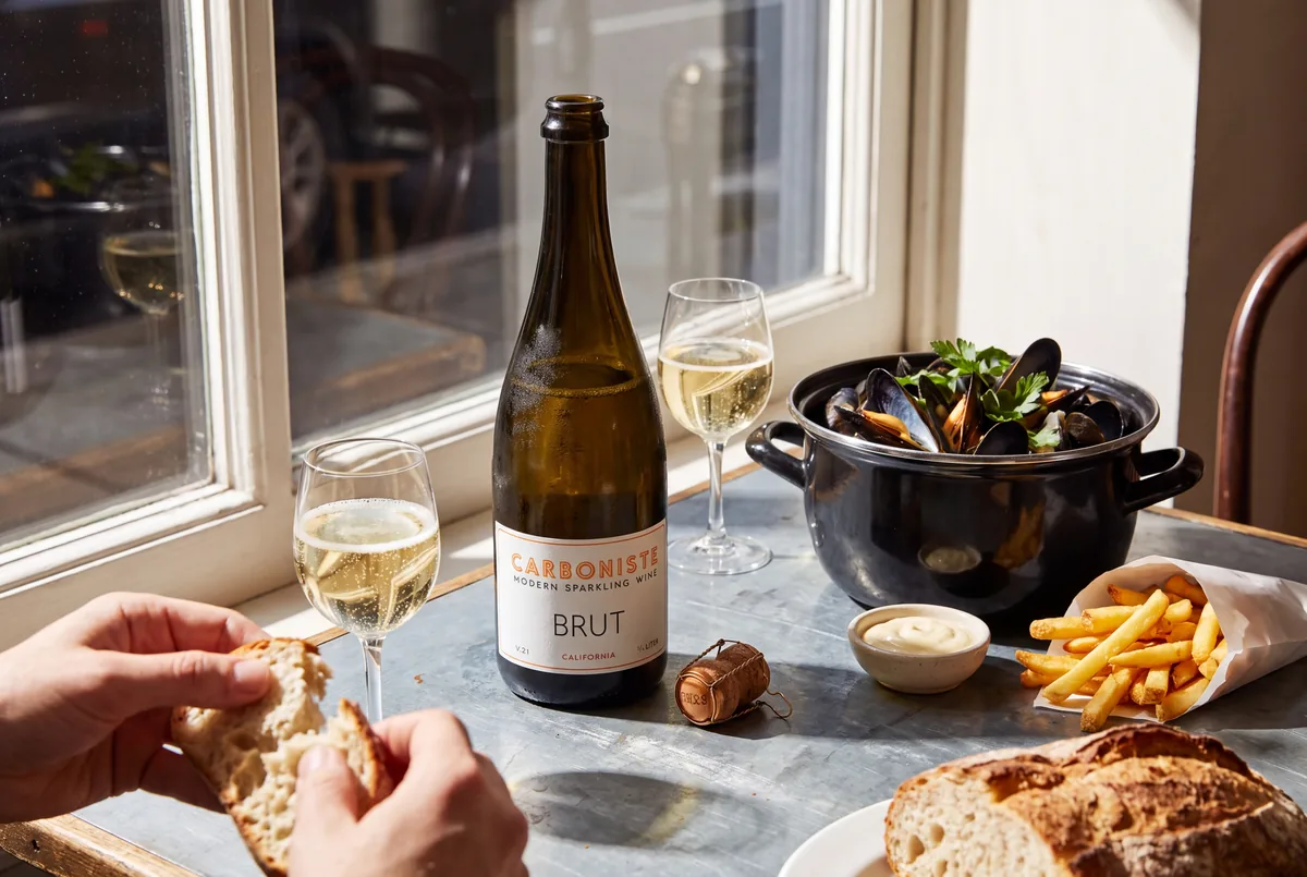 A bottle of Carboniste V.21 California Brut with its cork and cage beside it on a sunny bistro table with a pot of mussels, fries, aioli and two glasses of sparkling wine