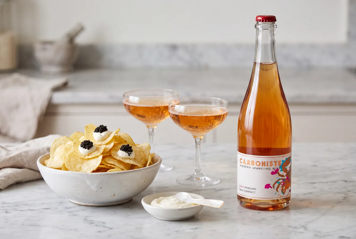 A bowl of plain potato chips topped with crème fraîche and caviar next to a bottle of Carboniste sparkling wine and two glasses